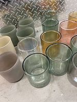 Photo showing assorted glass candle vessels in green, amber, pink, clear, yellow, and grey colors arranged on a metal surface.