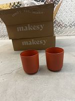 Two reddish-orange glass votive holders displayed side by side on a concrete floor, with two 'makesy' branded cardboard boxes in the background.