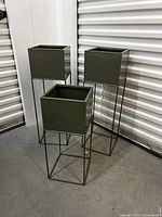 Three square olive green metal planters of different sizes on slim metal stands with geometric open bases, arranged together in a storage area.