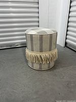 Full view of the round ottoman showing the overall plaid fabric, fringe trim, and cylindrical shape