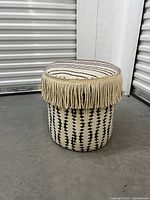 Full view of the round ottoman showing black geometric pattern on beige fabric and fringe detail under floral cushion top