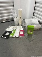 Overview of packaging items including stacks of white disposable cups, colored bags, white bowls with lids, some plastic clear packaging and a green box with window.