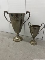 Full view of the two pewter trophies on a concrete floor against a white backdrop.
