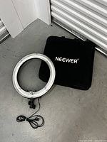 Neewer ring light with attached power cord and black carrying case placed on a concrete floor.