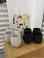 Front view of four white and four black stoneware mugs arranged on a wooden surface with the colorful GharyanKate Space Cookbook behind them.