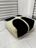 Full view of square tufted floor cushion with black and cream design on side, resting on white cloth.