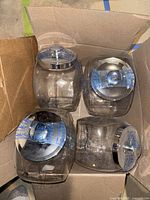 Four clear glass Anchor Hocking jars, square-shaped with rounded corners, silver lids, packed in original cardboard box.