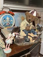 Porcelain teddy bear figurine seated, working with tools, alongside a stork figurine and kids alarm clock in the background.