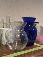 Photo showing a clear rounded vase with wide neck and a deep blue glass vase along with other glassware items.