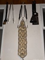 Photo shows six BBQ grilling tools hanging on hooks including a basting brush, tongs, two-prong fork, and three spatulas with black handles. Below the tools hangs a dried garlic art piece in a beige mesh netting, displaying multiple whole garlic bulbs.