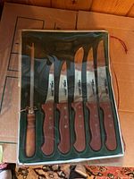 Photo showing five cutting knives with brown wooden handles and a sharpening steel in original green foam-lined box under a clear plastic cover.