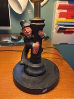 Close-up of the base of the art deco table lamp showing a figurine of a man in a black suit and top hat, holding the lamp pole and painted bottle detail. The base is black with some wear visible.