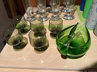 Mini green glasses arranged on a surface alongside the green swan container, showing the size and shape of the glasses and swan container.