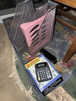 Lot items shown on a patterned rug next to a wooden table, including silver metal mesh magazine holders, pink plastic shelf, black stapler, and boxed calculator.
