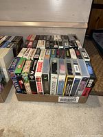 Photo showing one box of assorted VHS tapes including Julius Caesar, Triumph of the Spirit, Gone with the Wind, Lethal Weapon, 28 Mile, Men with Brooms, Maverick