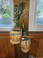 Two vintage Italian straw-covered Ruffino wine bottles with attached artificial decorative grape clusters displayed on a small wooden table by a window.