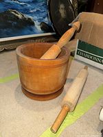 Photo showing heavy wooden mortar and pestle, mortar circular with carved lines, pestle fitting inside mortar, wooden roller on floor next to them.
