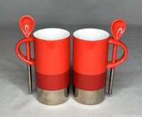 Two red and silver Starbucks espresso mugs each with a red spoon attached to the handle, side by side viewed from front angle.