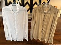 Front view of two men's linen long sleeve shirts, one white and one beige, both size XL, hanging on hangers.