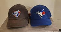 Two vintage Toronto Blue Jays baseball caps side by side on a wooden surface, one brown and one blue