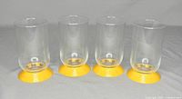 Four clear cylindrical drinking glasses each with a bright yellow circular base, shown in a front view on gray cloth background.