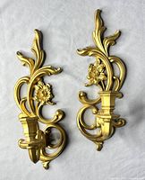 Pair of ornate gold vintage Syroco wall sconce candle holders shown lying on a white surface, displaying floral and leaf design details.