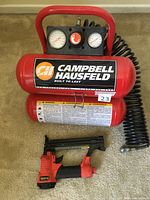 Full view of red Campbell Hausfeld air compressor with black gauge panel and coil air hose, with pneumatic nail gun placed in front