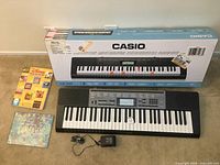 Overall view of Casio LK-265 keyboard on floor with power cord, two Disney sheet music books, and original box behind them.
