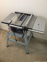 Front side view of the Delta ShopMaster table saw on metal stand showing the full table surface, saw blade slot with clear plastic guard, and stand leg design.