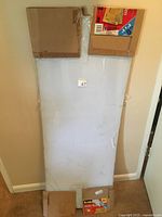 Front view of a large rectangular plexiglass sheet leaning against a wall with some bubble wrap and cardboard on and around it, showing size and condition.