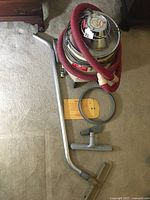 Industrial metal wet/dry vacuum with red hose, metal wand, wide vacuum head with bristles seen on carpet floor.