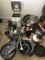 Wide shot of lot items on floor showing electric bread maker with open lid, coffee grinders, large bowls, and various cookware and utensils scattered.