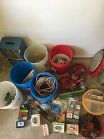 Wide view showing five buckets filled with various hardware parts and electrical wires, plus some small packaged items on floor and drawer units.