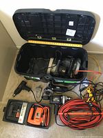 Overview of the open locking chest with power tools, including black reversible drill, angle grinder, shop light, extension cord coiled in front, and two circular saws.