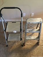 Front view of two aluminum step stools side by side: one is silver with two steps and black foam handle, the other has worn white paint and three steps.