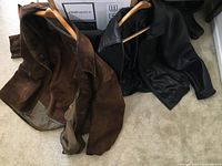 Two men's leather jackets on hangers, brown suede and black leather, displayed side by side on a carpet.