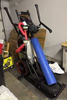 Front view of Bowflex Max Trainer M6 with attached red resistance tube and blue exercise mat leaning against foot pedal