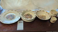 Photo of 2 bowls, 1 plate, 1 regular cup and 1 smaller cup showing Bunnykins storybook illustrations and bunny rim design.