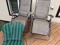 Two metal frame outdoor chaise lounge chairs with beige laced fabric seating and green striped cushions in front.