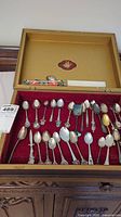 Wide shot of the open display box showing multiple collectible teaspoons arranged on red velvet lining, showcasing various handle designs and metals.