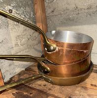 Two stacked copper saucepans with brass handles on wooden shelf