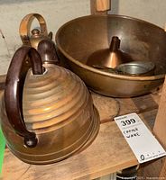 Photo of ribbed copper teapot with wooden handle and showing patina on body, placed on wooden surface with a large copper bowl, funnel, and measuring cups visible behind it.