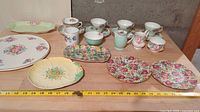 Full lot image showing assorted teacups, plates, trays, and cream pitchers arranged on a wooden surface with measurement tape for scale.