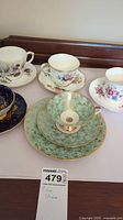 Five fine china teacups and saucers arranged on a white surface, various floral and gold designs visible.