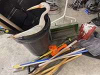 Photo showing black plastic garbage can, wooden handle tools, orange and black electric hedge trimmer, decorative bird figure, green spreader, and shovels.