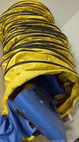 Yellow pet agility tunnel coiled with visible black reinforcement rings and dirt. Blue carrying bag is partially inside the tunnel, appearing worn and soiled.