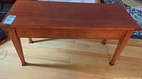 Full view of rectangular wooden piano bench with four legs on hardwood floor.