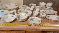 Full set of Royal Albert 'Forget Me Not' china with plates, bowls, cups, and saucers arranged on a shelf with a measuring tape in front showing approximate dimensions.