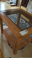 Angled view of the wood frame side table highlighting the glass top and cane shelf below, set on a patterned rug.