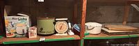 Wide view of shelf with multiple vintage kitchen appliances arranged including a rice cooker in box, Rival crock pot, kitchen scale, white Dutch oven, and warming plate with cutting board on top.